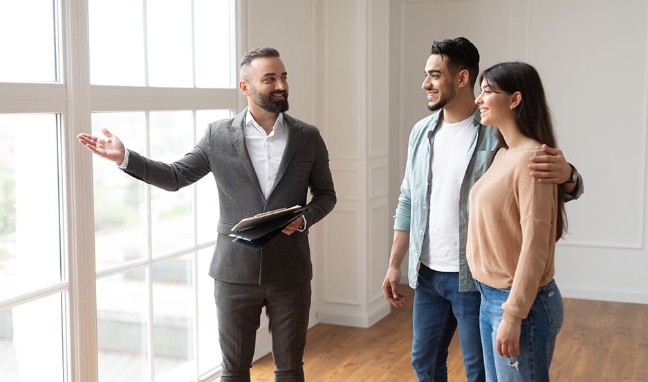 Portrait Of Male Realtor Or Architect Wearing Suit Showing View From New Empty House To Smiling Millennial Buyers, Pointing At Window. Husband Hugging Wife, Visiting Residential Building Choosing Home
